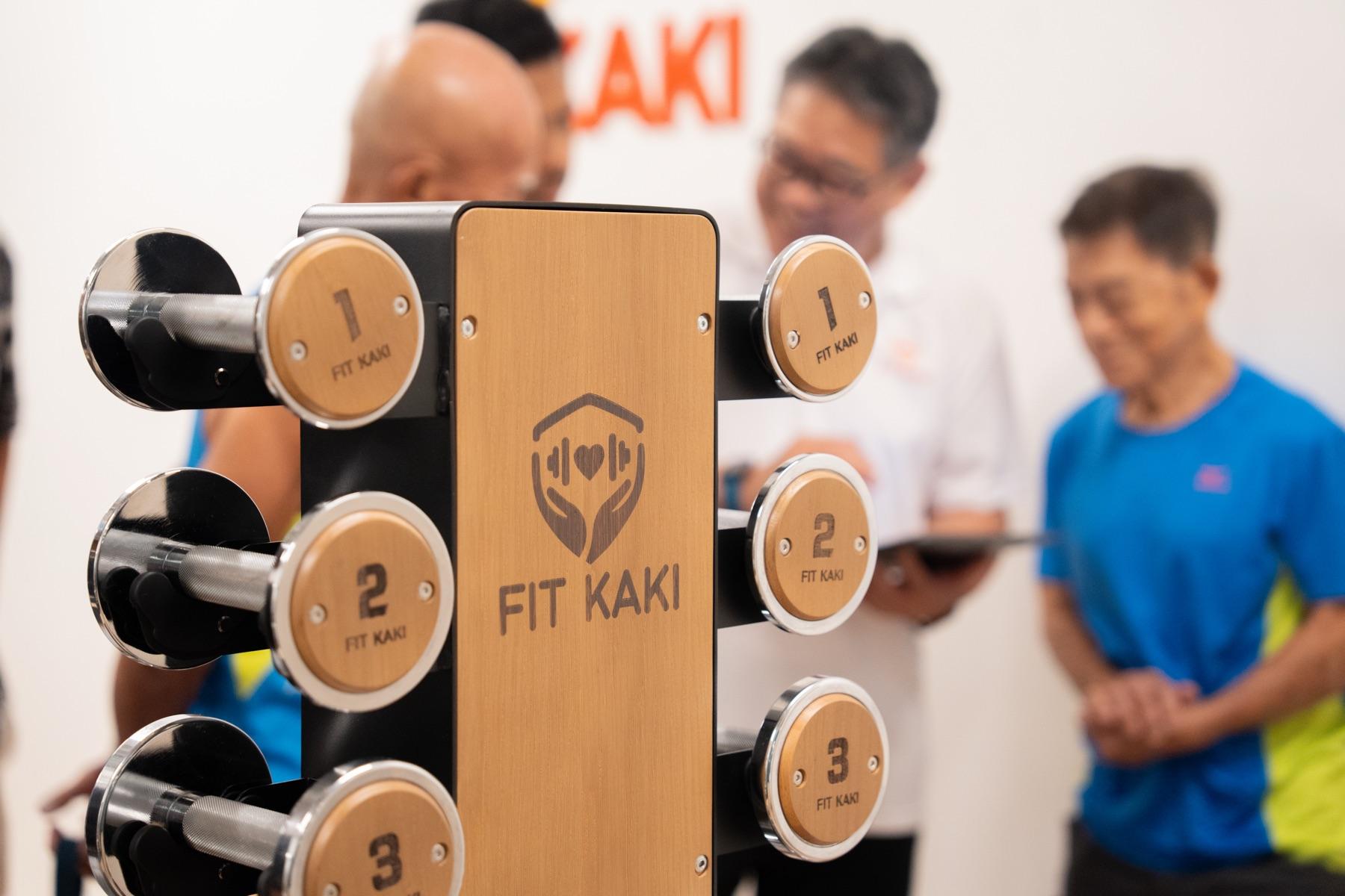 Inside the Fit Kaki Eunos studio: wooden dumbbell rack in front, coaches and seniors in a small session behind